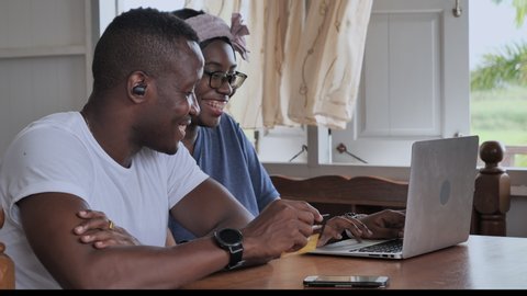 Black couple look happy with shopping online concept at home. They are sitting in living room, using smartphone, laptop, and entering credit card details on laptop, Quarantine at home for social dista