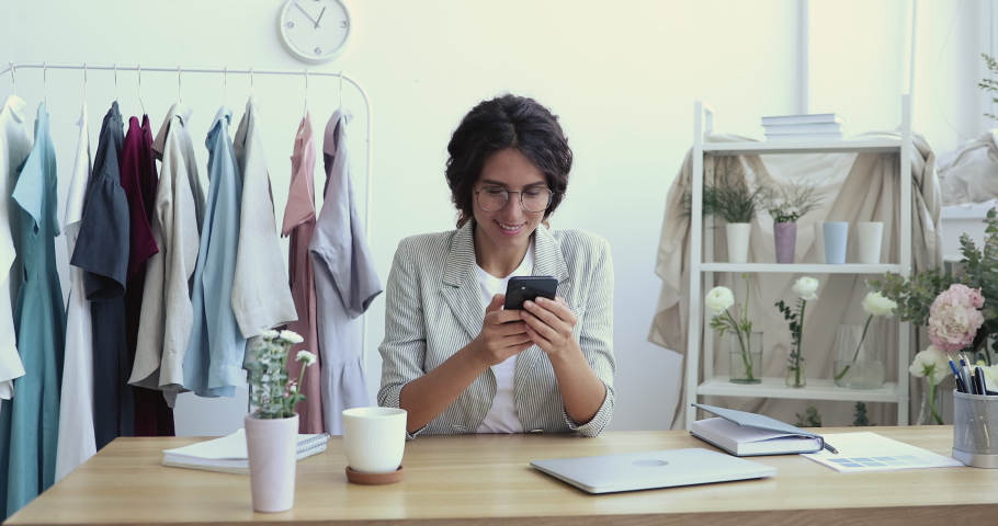 Attractive young female entrepreneur in eyewear sitting at table, using smartphone, communicating with clients or involved in doing marketing strategy in social networks online in modern boutique. Royalty-Free Stock Footage #1057830001