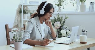 Smiling beautiful focused young female creative decorator florist in wireless earphones looking at laptop screen, watching educational lecture online seminar webinar, writing notes at workplace.