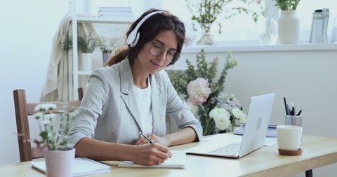 Smiling beautiful focused young female creative decorator florist in wireless earphones looking at laptop screen, watching educational lecture online seminar webinar, writing notes at workplace.