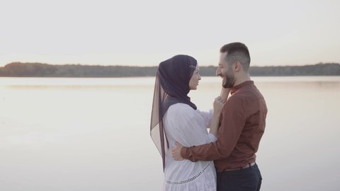 Muslim love story video in 4k. Mixed couple smiles and walks near lake, than turns around embraces and looks at sunset . Woman weared in hijab looks to her man. Advert for on-line dating agency