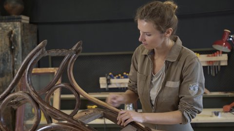 A working female carpenter peels off paint from rocking chair legs with a spatula, restorer in a workshop, handheld shot. Restoration of wooden rocking chair in a workshop. 