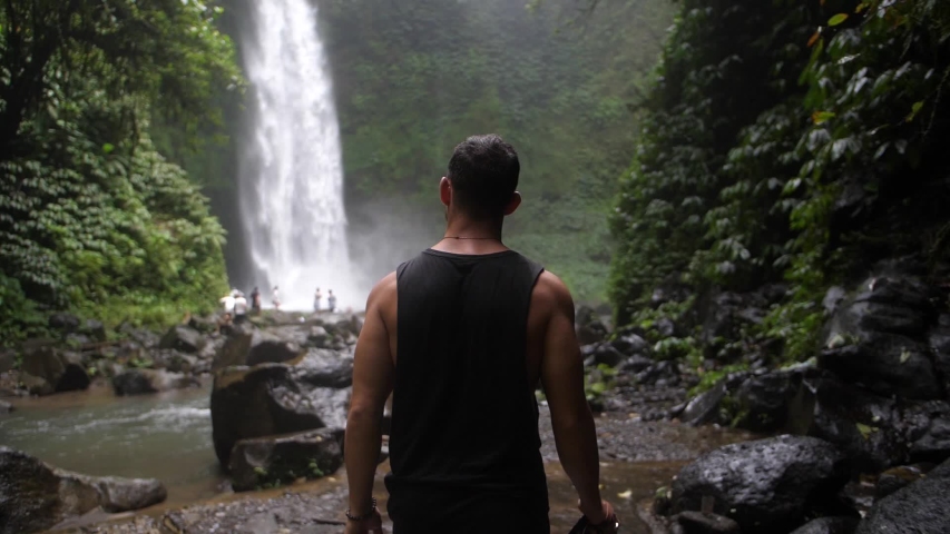 A young, fit man is walking by himself towards a waterfall in the depth of the Bali jungle. Holding a big and professional DSLR camera to take photos and videos. Slow motion. | Shutterstock HD Video #1057840573