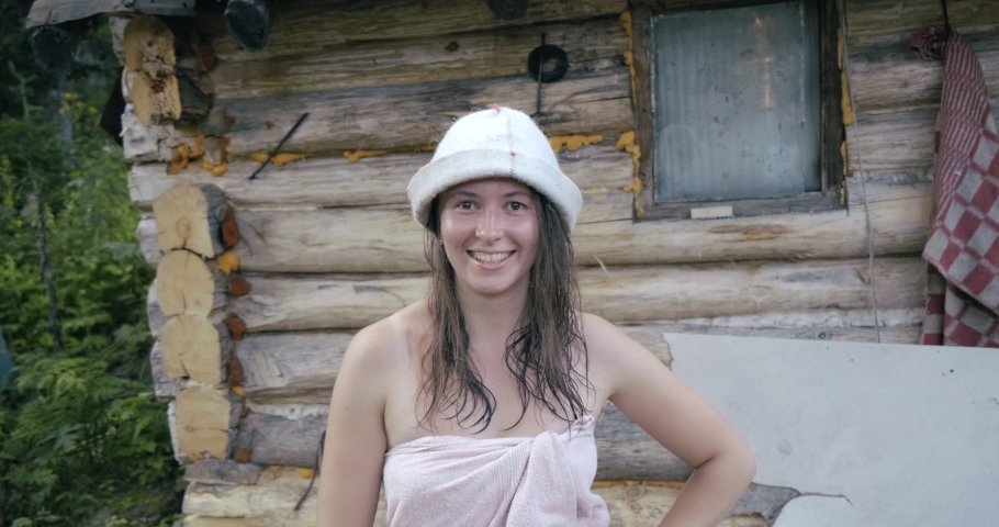 Pretty woman Stands by Log Building. Wrapped in Towel. Came out of Sauna. Village. Forest. Wilderness. Taiga. Sauna Cap on Woman