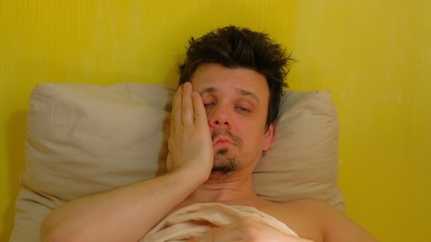 Sleepy tired bearded man with hangover wakes up in bed looking at camera, portrait closeup. Young man with headache in the morning after waking up. Tired sick guy. Yellow wallpaper background.