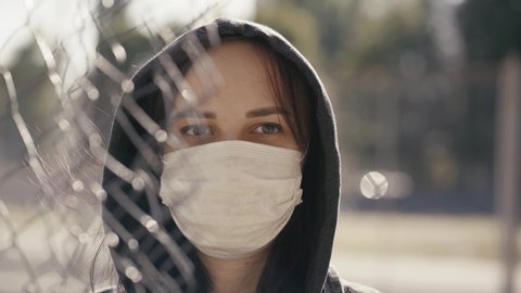 Portrait of young woman in hood and medical mask in sunny weather. Close up of adult female through lattice, protecting yourself from diseases in period coronavirus epidemic infection.