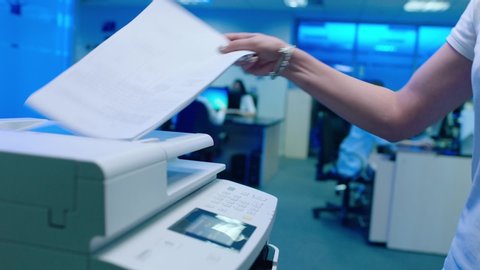 Woman using printer or scanner in office . Office worker use print machine on blurred office background with workers and computers . Hand take papers and press buttons on multifunctional copier .