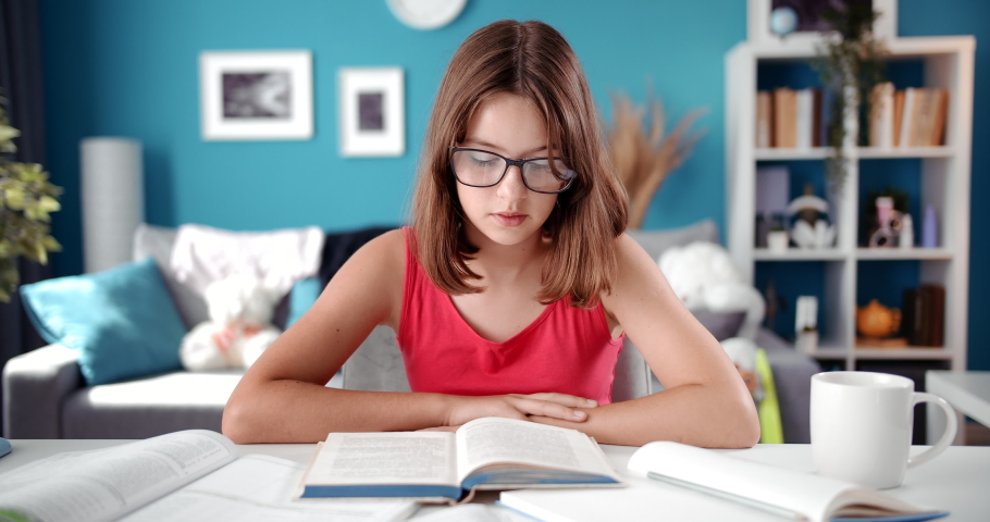 Front view of schoolgirl in eyeglasses sitting at table and reading interesting book. Concept of self education and kids activity during quarantine.
