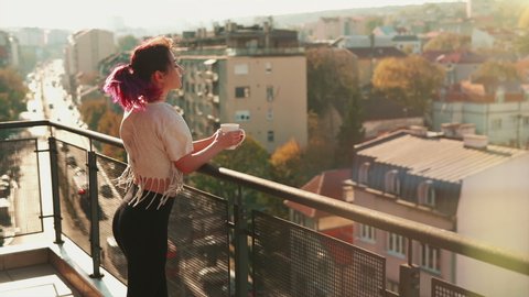 Gorgeous young woman with purple hair stays on balcony holds cup of hot coffee or tea watching a beautiful urban view and then close eyes and enjoy relax breathing fresh freezing air of spring