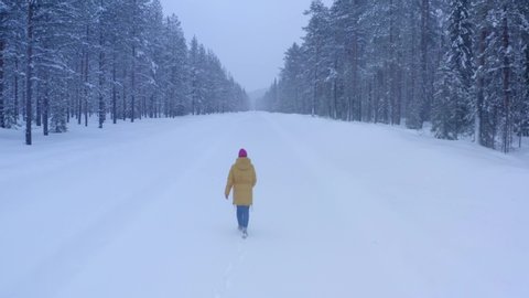 Bird's eye view, female tourist in yellow warm jacket enjoying winter in north country while running on snowy road surrounded by coniferous forest. Wanderlust in journey, freedom in life, having fun: film stockowy