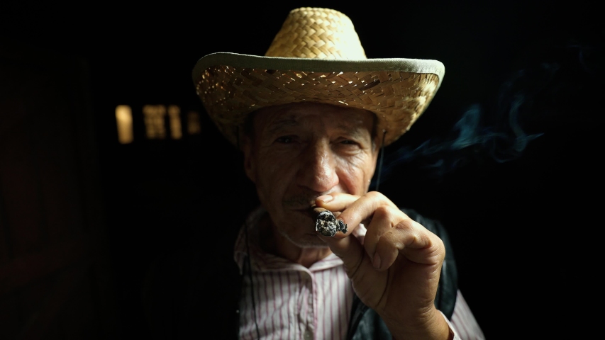 The man smokes a cigar. An elderly farmer in a straw hat smokes a cigar outside the ranch. Thick tobacco smoke. Confident Senor reflects in cigarette smoke. An old cowboy in a leather vest | Shutterstock HD Video #1057869148