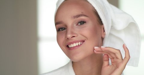Close-up of Woman with Smooth Healthy Skin, Looking in Mirror, Touching her Face Gently, Smiling. Portrait of Girl Model in White Bathrobe and Towel Posing for Camera. Skincare Concept.