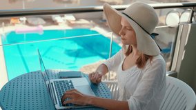 Woman Talks Online using Laptop in Hotel with Swimming Pool on Summer Holiday or Vacation. Lady in Straw Hat makes Video Call and Greets Friend. 4K Medium Orbit Handheld Shot