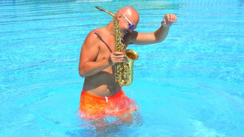 Blurred defocused male musician saxophonist with naked torso in orange swimming trunks dancing, having fun in pool, playing golden alt saxophone on musical instrument. Summer party. Slow motion video.
