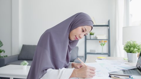 Beautiful Asia muslim lady in headscarf casual wear using laptop in living room at house. Remotely working from home, new normal lifestyle, social distancing, quarantine for corona virus prevention.