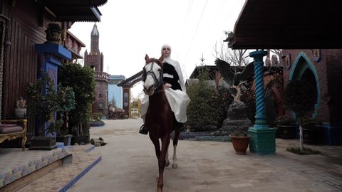 attractive lady in traditional Arabic costume with hijab rides brown horse along yard of oriental restaurant slow motion