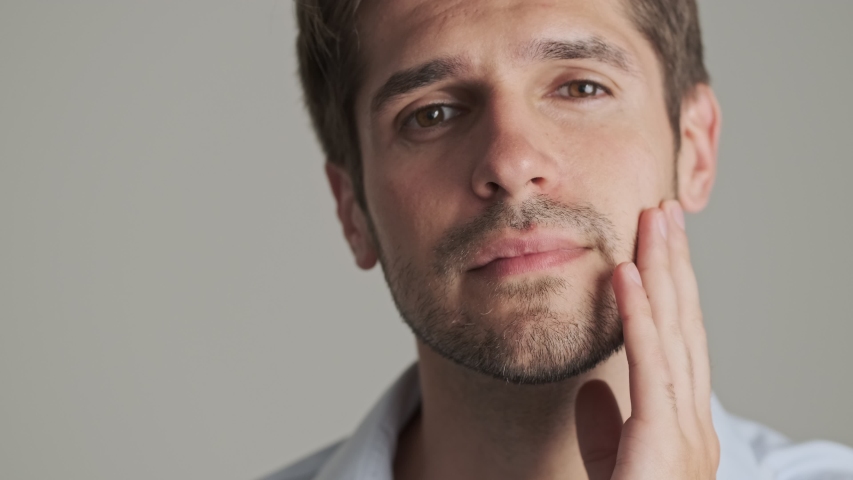 A close-up view of a pleased unshaven young man is looking at himself in the mirror standing isolated over white background. Royalty-Free Stock Footage #1057887601