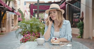 Positive smiling woman is talking with friend by the phone while sitting in the small street cafe. Beautiful traveler enjoying coffee at bakery on street of old European town. Wireless connection 4K