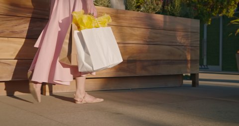 Woman in pink dress with shopping bags walking in a city at sunset. Slow motion of female in stylish outfit is walking by the outdoor shopping mall on sunny summer evening. Malibu, California, USA
