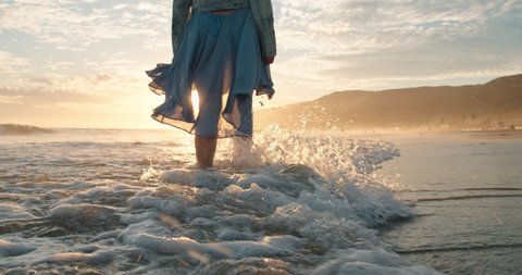Slow motion woman walking barefoot in ocean waves in golden sunset light. Cinematic 4K of happy female traveler enjoying her summer vacation in Malibu, California, USA. Woman in beautiful waving dress