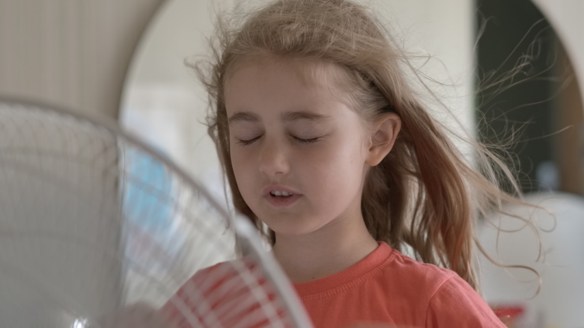 Girl Standing in Front of Fan. Child Enjoying Cool Wind From Electric Fan at Home at Summer Vacation. Suffer From Heat High Temperature in Front of Ventilator Cooling Herself With Electric Fan-Cooler. Royalty-Free Stock Footage #1057890244