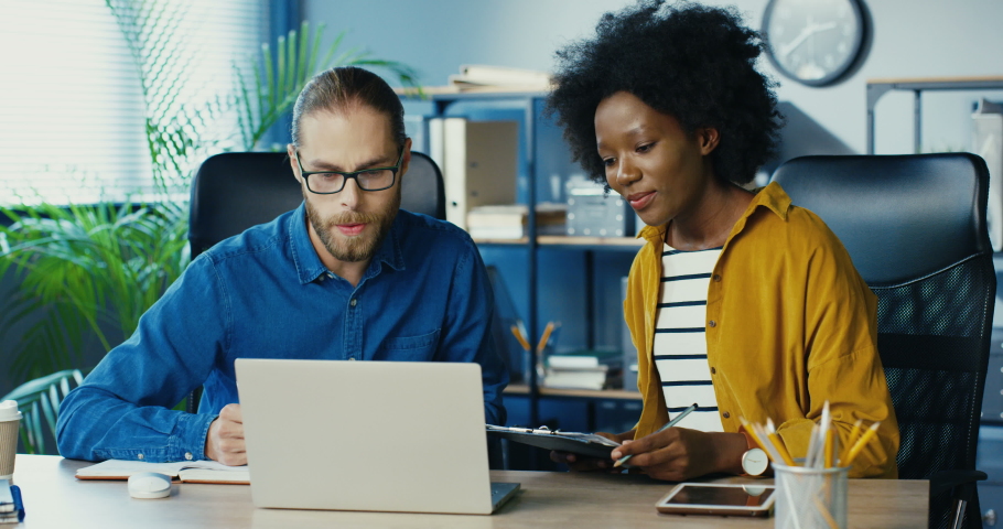 Mixed-race office workers having online video presentation on laptop at office. Pretty African American girl showing graphs to webcam. Handsome Caucasian man in glasses explains diagrams on video call | Shutterstock HD Video #1057898980
