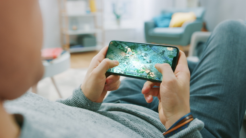 Point of View Shot Man Playing Mockup- MOBA RPG Game on his Smartphone while resting at Home on a Couch. Cool Game with Modern Graphics and Fun Gameplay. Cyber Gaming Royalty-Free Stock Footage #1057900351