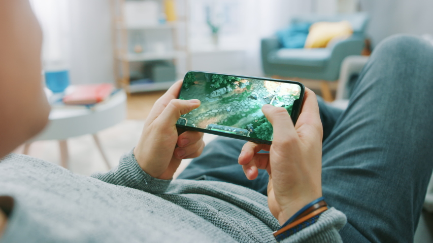 Point of View Shot Man Playing Mockup- MOBA RPG Game on his Smartphone while resting at Home on a Couch. Cool Game with Modern Graphics and Fun Gameplay. Cyber Gaming | Shutterstock HD Video #1057900351