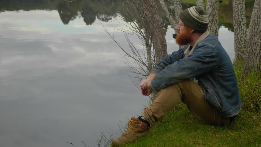 A mid shot of a bearded ginger mountain man smoking a tobacco pipe while sitting by a calm lake.