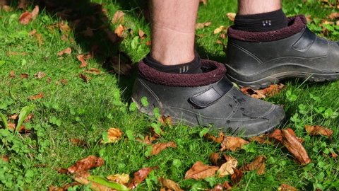 Slow motion. a young man in dirty black galoshes with bare hairy legs walks on a lawn of green grass, lit by bright sunlight.