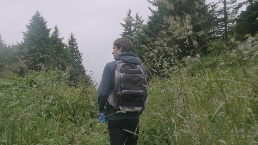 Young white caucasian male hiking walking with backpack through nature trail passing tall grass plants in beautiful lush green forest trees tracking shot overcast day oregon handheld shot slow motion