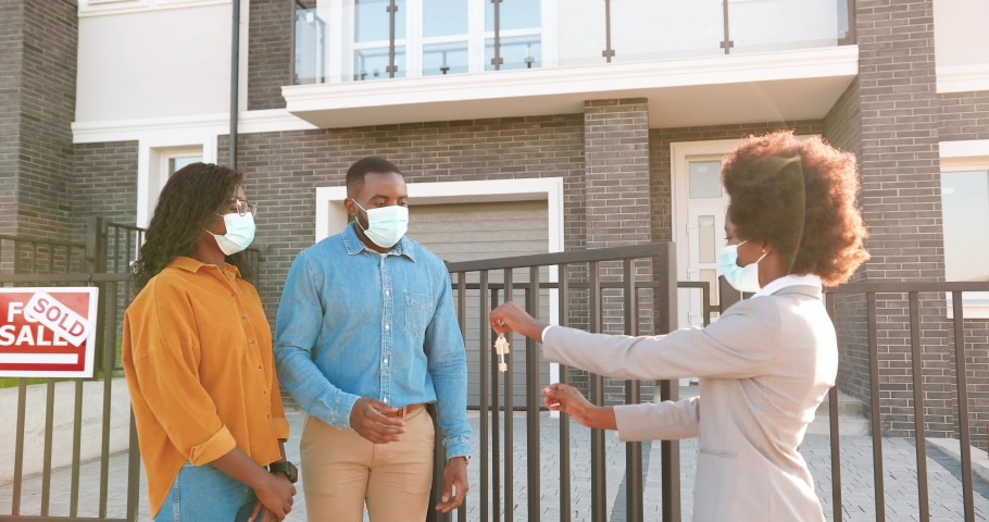 Female real-estate selling house to African American happy couple and passing keys. Outdoor. Cheerful just-married man and woman in medical masks hugging when buying home at outskirt. Covid-19 concept Royalty-Free Stock Footage #1057902814
