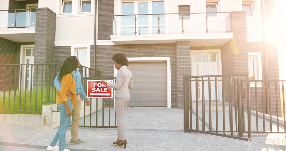 African American young female real-estate agent in business style with documents in hands showing house to happy just-married couple. Outdoors. Looking and buying new home. Suburb neighborhood. Royalty-Free Stock Footage #1057902844