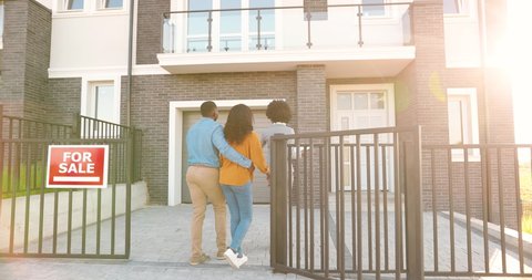 African American young female real-estate agent in business style with documents in hands showing house to happy just-married couple. Outdoors. Looking and buying new home. Suburb neighborhood.