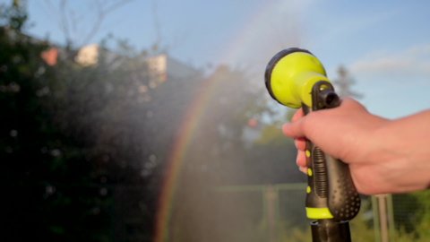 Close up female hand holding water hose and watering lawn or plants in back yard. Gardener woman with sprinkler in garden at evening sunset. Rainbow is visible in sun.