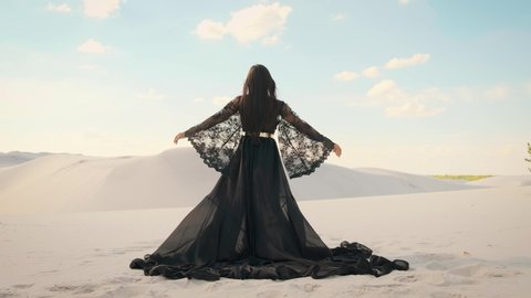 Beauty woman, rear view. Queen in black clothes dancing in desert. Girl fashion model. long silk arab dress. Back of luxury elegant goddess. backdrop white sand, blue sky. Mysterious silhouette queen