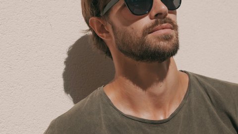 Portrait of handsome stylish hipster lambersexual model. Man dressed in T-shirt. Fashion male posing in the street near white wall outdoors in sunglasses. He looking at camera and grins