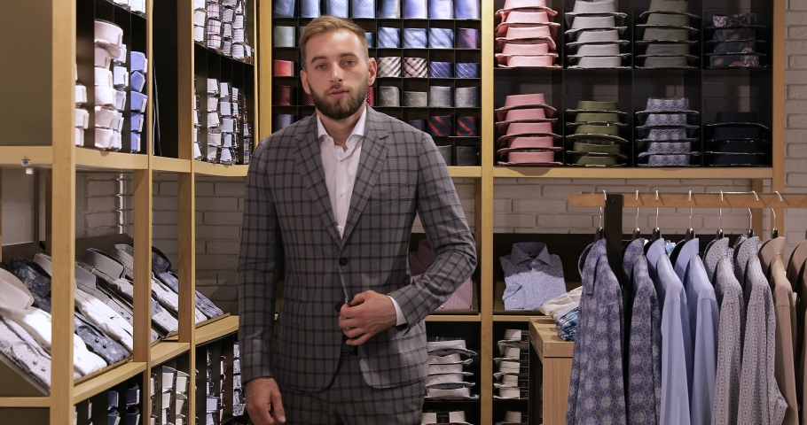 Front view slow motion general shot of a man in gray business clothes standing in a clothing store looking straight at the camera straightening his hair and smiling