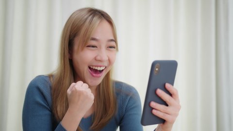 Excited Asian Woman having good news on phone smile and enjoy success on phone. Joyful Girl reading good news making yes gesture celebrating good news while sitting in living room at home