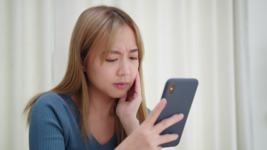 Upset Asian woman looking at smartphone screen feels sad and disappointed with bad news messages on phone. Unhappy Asia Female Receive bad news while sitting in living room at home Royalty-Free Stock Footage #1057914628