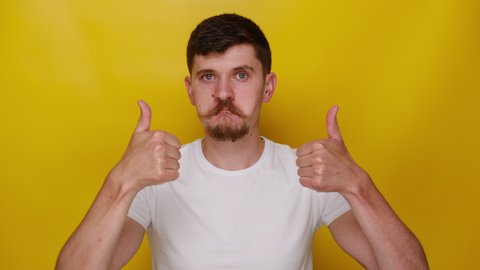 Young man winking and vigorously showing double thumbs up, like gesture, demonstrating approval, feeling enthusiastic satisfied with excellent result, wears t-shirt, isolated on yellow background
