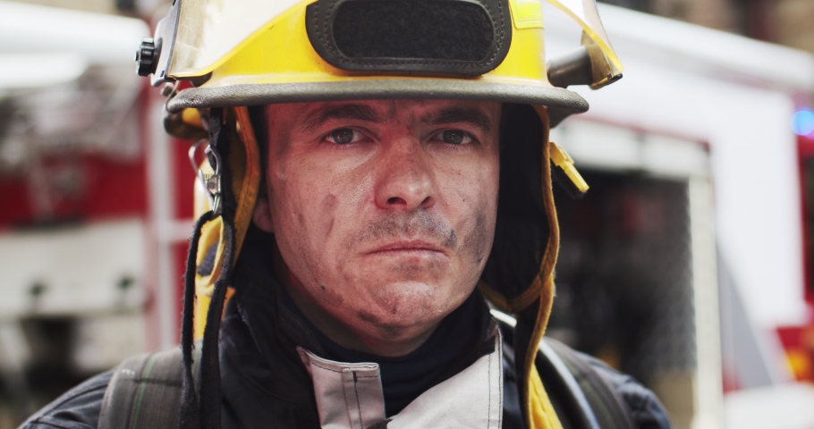 Close up portrait of strong serious fireman in helmet and gull equipment standing next to car with flashing lights on and looking into camera. Concept of saving lives, heroic profession, fire safety