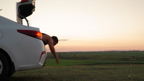 A white car pulls up against the backdrop of a beautiful sunset and an athletic man out of the trunk and walking on hands. Handstand. Sports humor.