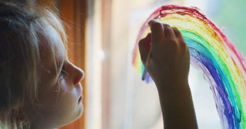 Authentic shot of a cute little creative happy smiling preschooler girl painter is drawing a rainbow on a glass of a window in a kitchen at home. Concept of happiness, arts, childhood, covid free