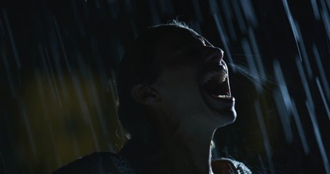 Cinematic shot of an young carefree happy woman with loose hair in white dress is screaming of freedom and enjoying the rain showering her with wide arms open in a hot summer in a city at night.