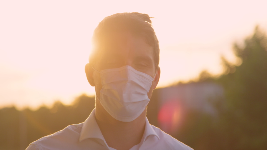 SLOW MOTION, CLOSE UP, PORTRAIT, HANDHELD, LENS FLARE, DOF: Young Caucasian man takes off surgical facemasks at golden sunset. Man is relieved to remove mask off his face during covid-19 pandemic. Royalty-Free Stock Footage #1057931776