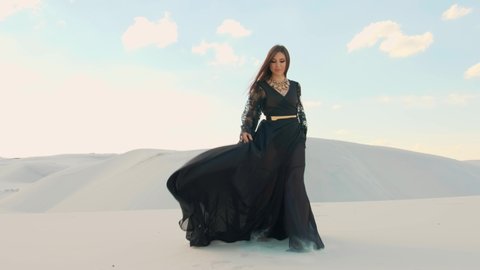 Young woman in black stylish dress with long sleeves walks in the desert. Arabian beauty princess. Fabric and hair are blown in the in slow motion. Arab emirates nature, white sands, summer desert