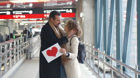 Beautiful woman holding board with red heart sign meeting husband in airport terminal. Happy couple embracing and smiling after long separation at railway station