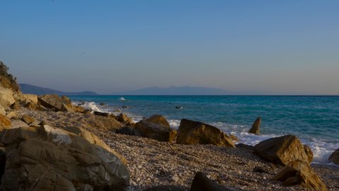 Sea waves splashing on rocks of peaceful beach with pebbles, seascape with Corfu island in background