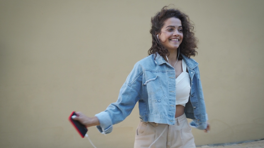 Active happy smiling latin hispanic teen young woman dancing in slow motion while listening energetic music on her smartphone in earphones. Young woman having fun outdoors in front of background wall. | Shutterstock HD Video #1057941010
